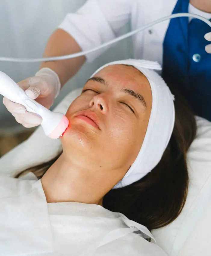 A woman with a headband lies on a treatment table, receiving a laser therapy treatment on her chin from a person wearing gloves.