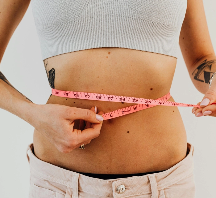 A person wearing a grey crop top and beige pants measures their waist with a pink measuring tape.