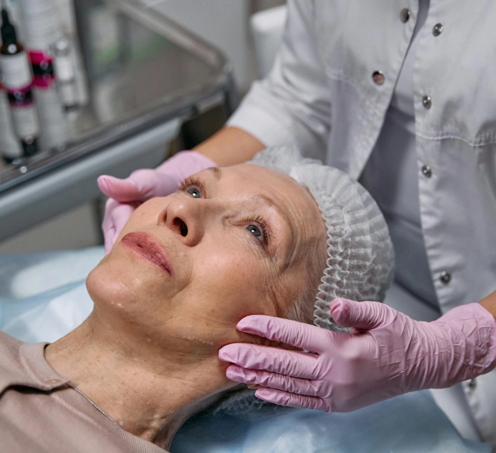 A woman with a medical cap lies on a clinic bed while a professional wearing pink gloves examines her face.