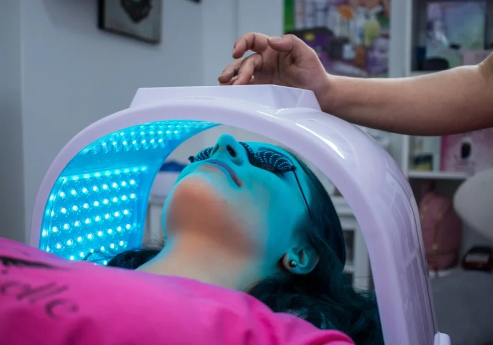 A person lies under a blue LED light therapy device with goggles on, while someone adjusts the machine above their head in a clinical setting.