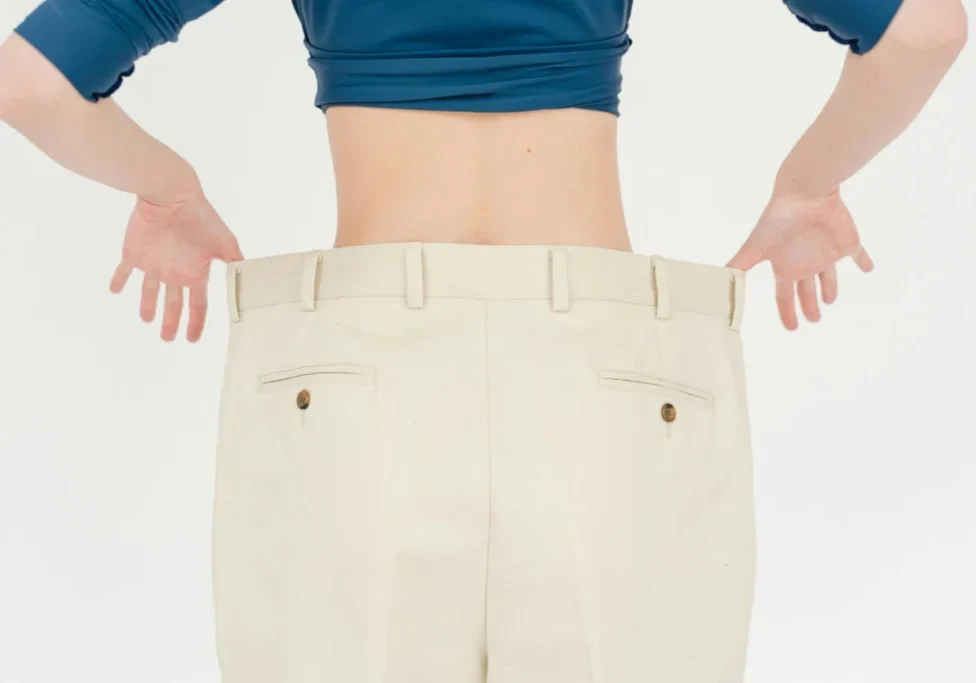 Person wearing oversized beige pants and a blue cropped top, holding the pants waistband out to show their large size. The image is taken from behind.