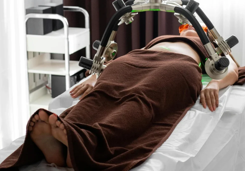 A person lies on a treatment table covered with a brown blanket, undergoing a laser treatment on their abdomen with a large, multi-armed machine in a clinical setting.